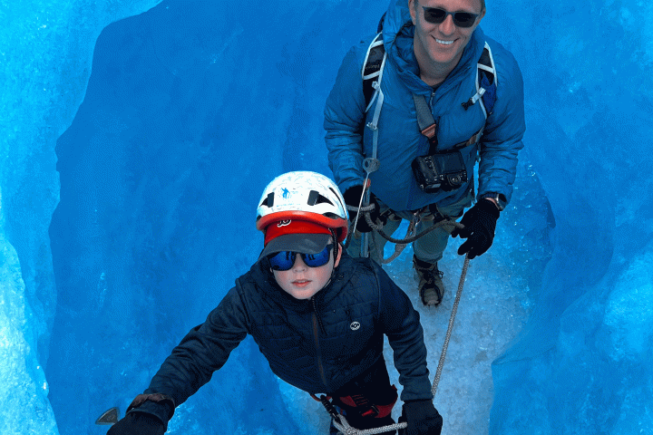 Two people in climbing gear stand inside a vibrant blue ice cave, looking up and smiling.