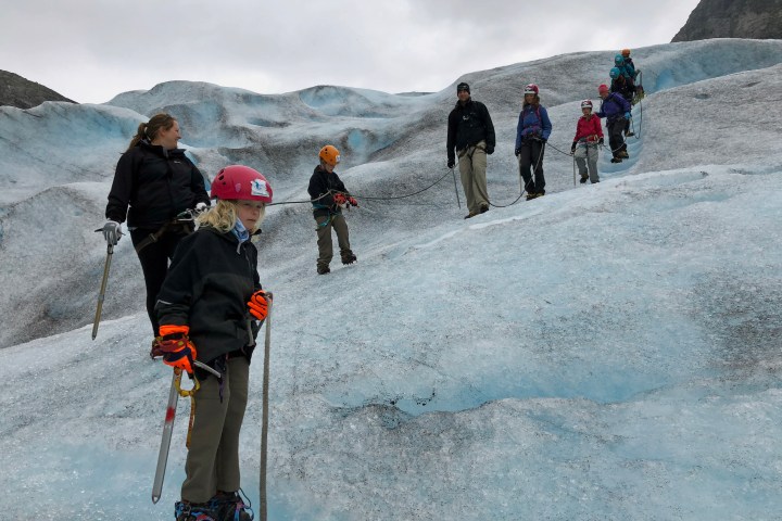People hiking on a glacier with climbing gear and helmets under a cloudy sky.
