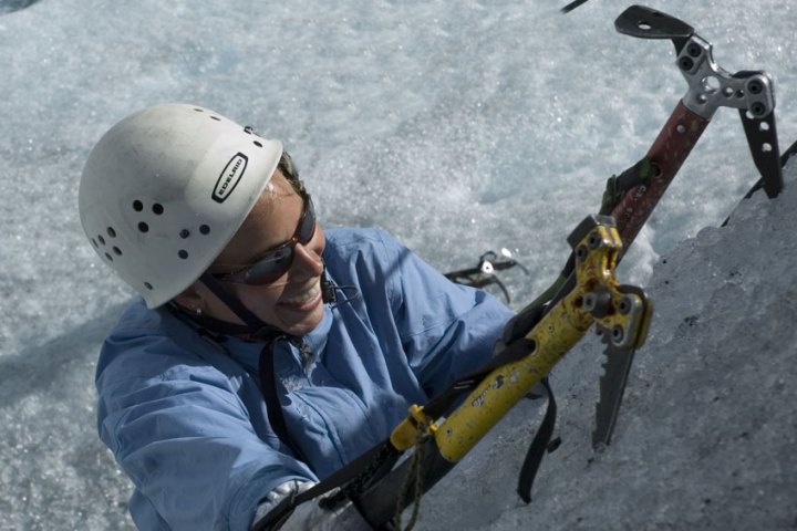 Climber with helmet and sunglasses scaling icy surface using ice axes.