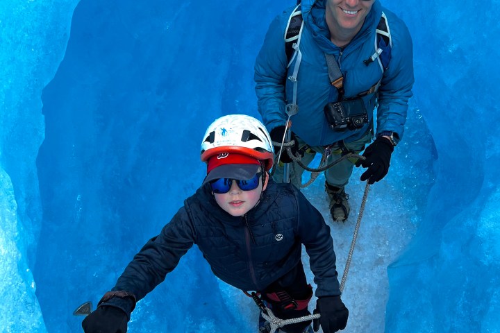 Two climbers in gear inside blue ice crevice, wearing helmets and sunglasses, holding climbing tools.